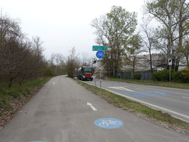 Asphaltierter Radweg mit entsprechender Beschilderung, daneben eine Straße ohne Gehsteig, jeweils in einer engen Kurve, auf der Straße fährt gerade ein LKW