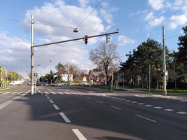 Straßenraum einer Kreuzung, in Blickrichtung ein eigener Straßenbahngleiskörper, daneben drei KFZ-Fahrspuren in eine Fahrtrichtung aber verschiedene Relationen, wiederum daneben ein Radweg und ein Gehsteig. Bei der Folgekreuzung zweigt eine Straße nach rechts ab, entlang der schließlich, inzwischen weit im Hintergrund, sich eine Bushaltestelle befindet. Diese wurde zur besseren Sichtbarkeit im Foto nachträglich rot eingekreist