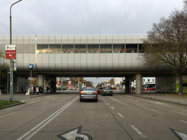 Straße unterquert eine in Hochlage liegende U-Bahn-Station, in Blickrichtung zwei KFZ-Fahrspuren, daneben eine Haltestellenspur für Busse mit Fahrrad-Mehrzweckstreifen, in der Gegenrichtung eine Fahrspur, daneben eine Haltestellenspur für Busse mit Fahrrad-Mehrzweckstreifen